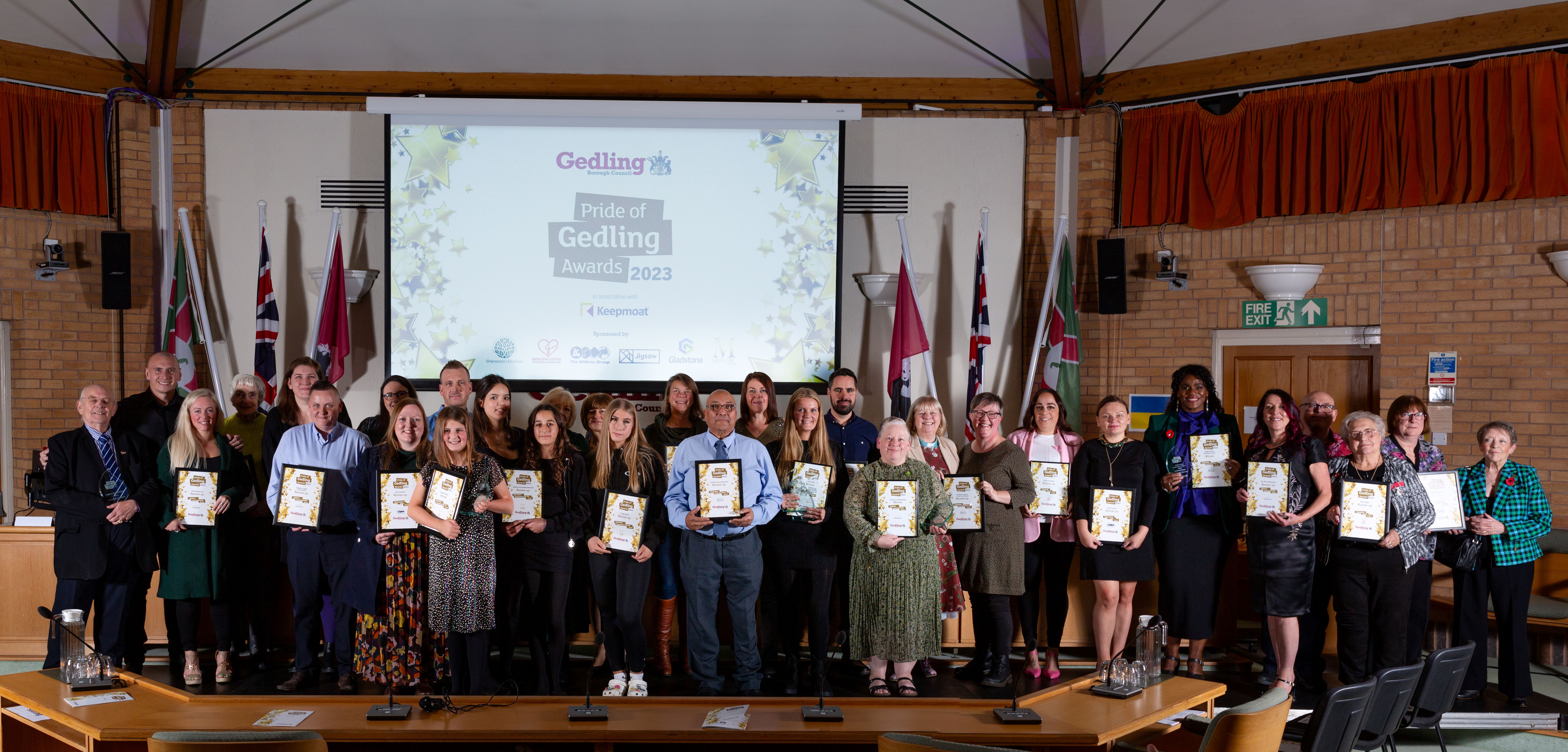 A large group of people stood in a row holding awards and certificates for the Pride of Gedling Awards 2023. The group are stood in front of a large projector screen which has text on it reading “Pride of Gedling Awards 2023”